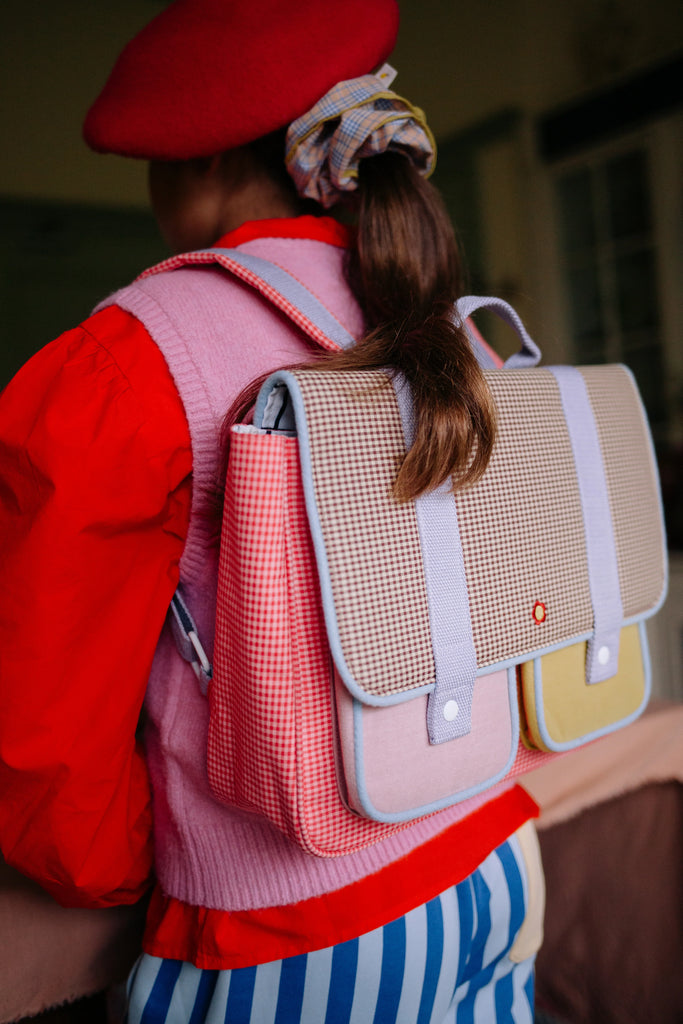 A person wearing a red beret, red shirt, pink vest, and blue-striped pants has the Sticky Lemon School Bag | Lemon Tree | Gingham Flower + Rose—crafted from recycled PET bottles and featuring a pastel gingham design—slung over their shoulder. Their hair is tied with a plaid scrunchie.
