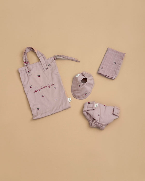 The Konges Sloejd Doll Nursery Set in Cherry Blush includes four mauve, organic cotton doll accessories—a tote bag with text, bandana bib, bloomers, and folded cloth—each with a charming small cherry print on a beige background.