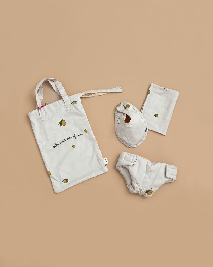 The Konges Sloejd Doll Nursery Set - Lemon features white organic cotton doll accessories with small yellow lemon prints: a bib, a bag, bloomers, and a folded cloth, all shown on a beige background.