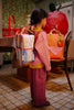 A young child with braided hair stands indoors wearing a pink sweater, red checkered pants, and the Sticky Lemon Backpack Small | Stripes | Cloud Blue + Twiggy Beige. Colorful bags and playful decor, including an apple-shaped object, are in the background.