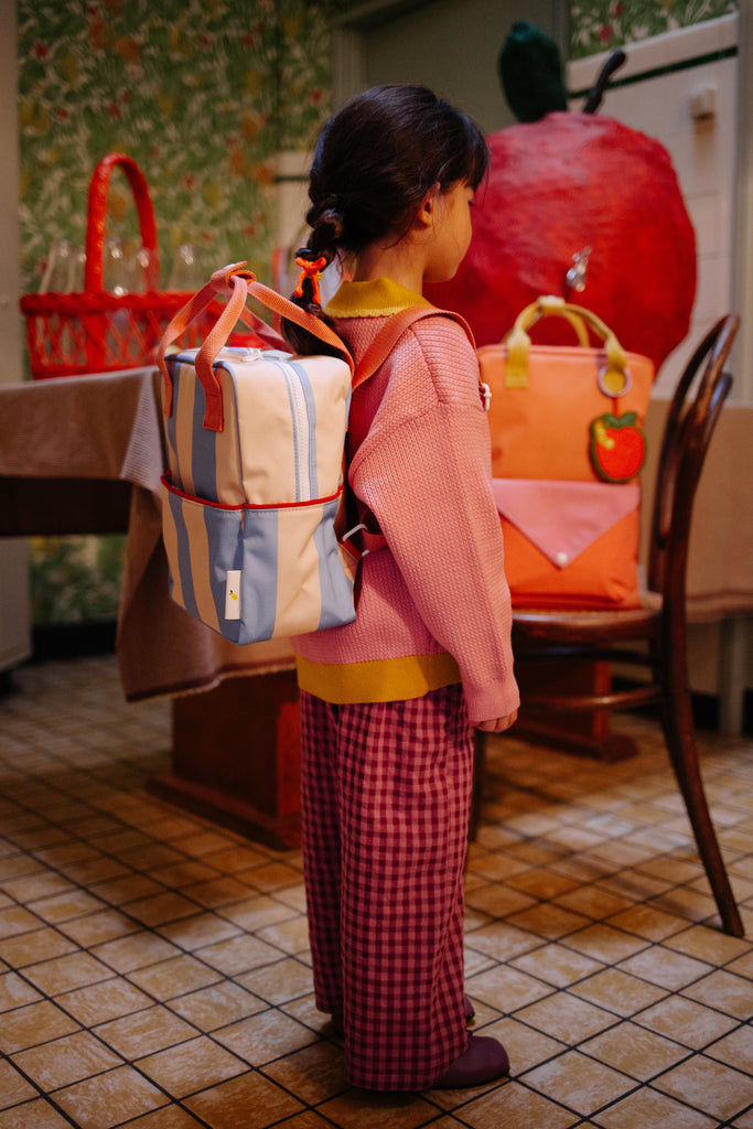 A young child with braided hair stands indoors wearing a pink sweater, red checkered pants, and the Sticky Lemon Backpack Small | Stripes | Cloud Blue + Twiggy Beige. Colorful bags and playful decor, including an apple-shaped object, are in the background.