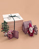 The Maileg Gift Box: Santa’s Christmas Wish features a Christmas Mouse in red, a mini tree, a gingerbread-style toy house, a tiny doll in a tin bed, gray pouch, and large wooden gift box with black ribbon arranged on a beige surface.