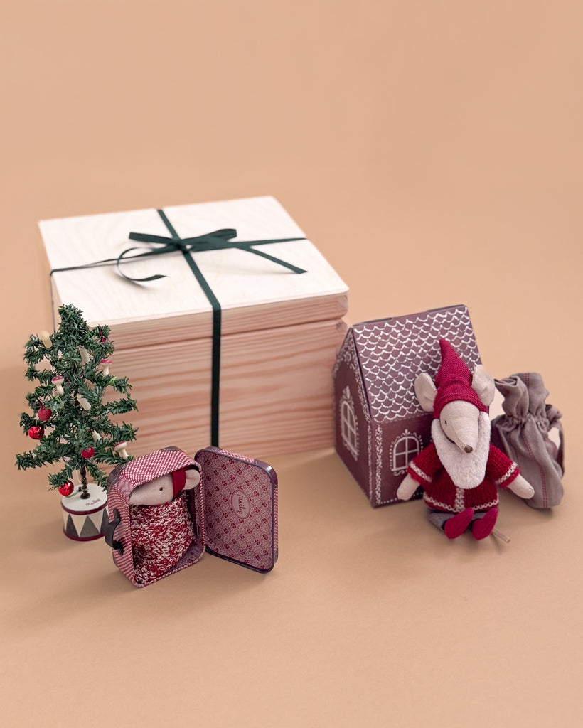The Maileg Gift Box: Santa’s Christmas Wish features a Christmas Mouse in red, a mini tree, a gingerbread-style toy house, a tiny doll in a tin bed, gray pouch, and large wooden gift box with black ribbon arranged on a beige surface.