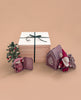 The Maileg Gift Box: Santa’s Christmas Wish, tied with a green ribbon, is displayed beside a small decorated Christmas tree, a Maileg Christmas Mouse, Mouse Gingerbread House, and patterned fabric pouches on a beige background.