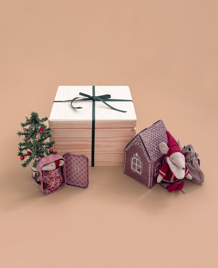 The Maileg Gift Box: Santa’s Christmas Wish, tied with a green ribbon, is displayed beside a small decorated Christmas tree, a Maileg Christmas Mouse, Mouse Gingerbread House, and patterned fabric pouches on a beige background.