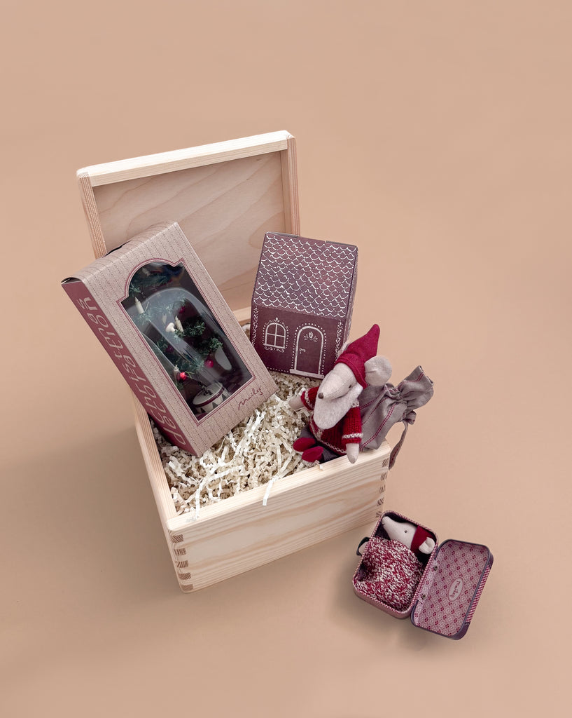 The Maileg Gift Box: Santa’s Christmas Wish features an open wooden box with paper houses, a small Maileg Christmas Mouse in a red outfit, shredded paper filling, and two small tins—one hiding another tiny fabric mouse—set against a beige background.