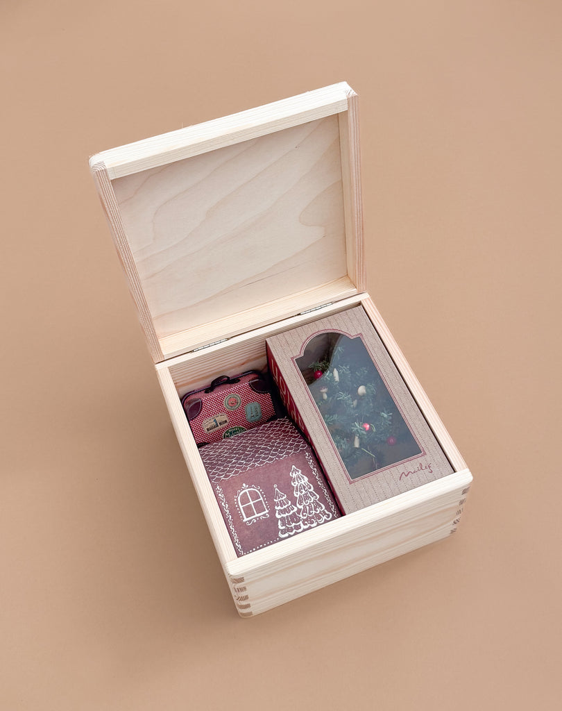 A light wooden box with an open lid displays the Maileg Gift Box: Santa’s Christmas Wish, featuring gifts like a burgundy camera, a festive notebook, and a Maileg Christmas Mouse in a windowed box against a beige background.