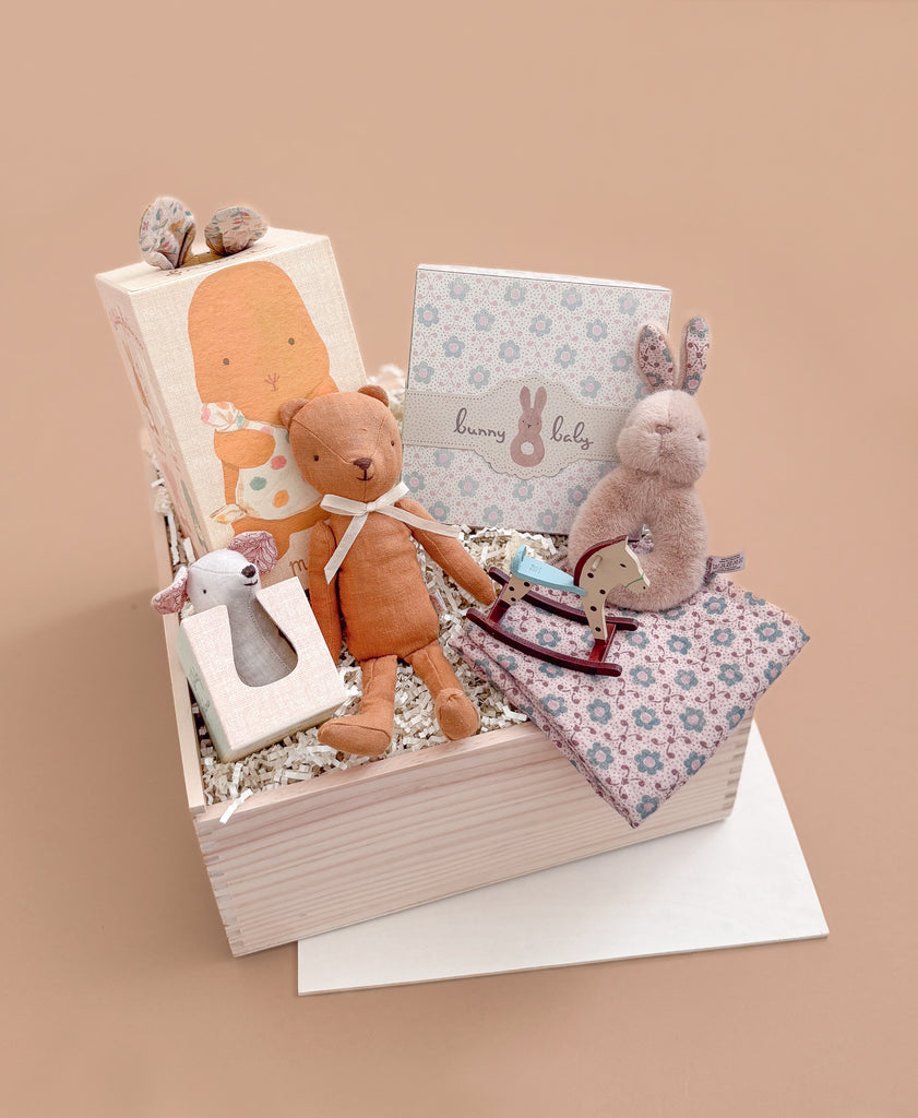 The Maileg Gift Box: Little One’s First Friends includes a Bunny Rattle, Lullaby Friends stuffed animals, a floral blanket, “bunny baby” card, and wooden rocking horse, all in pastel packaging with white shredded paper on a beige background.
