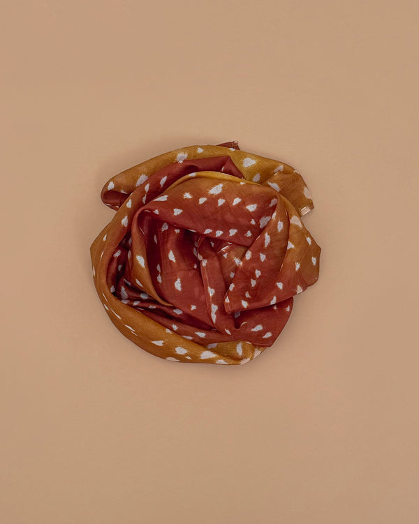 A rust and mustard-colored scarf with white geometric patterns, inspired by Sarah's Silks Animal Print Playsilk - Fawn, is coiled in a circular shape on a beige background.