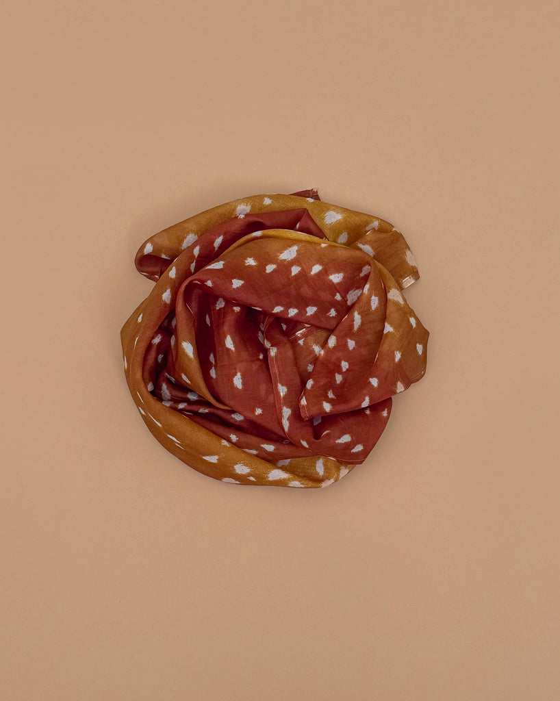 A rust and mustard-colored scarf with white geometric patterns, inspired by Sarah's Silks Animal Print Playsilk - Fawn, is coiled in a circular shape on a beige background.