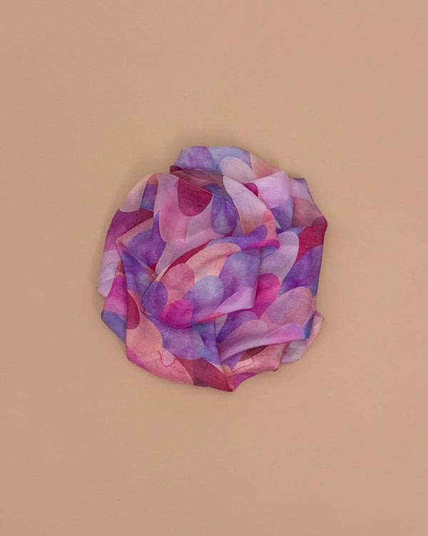 Sarah's Silks Magical Creatures Playsilk - Pink Unicorn, made of pure silk with overlapping purple, pink, and lavender shapes, is artfully arranged in a circular, flower-like design on a plain beige background.