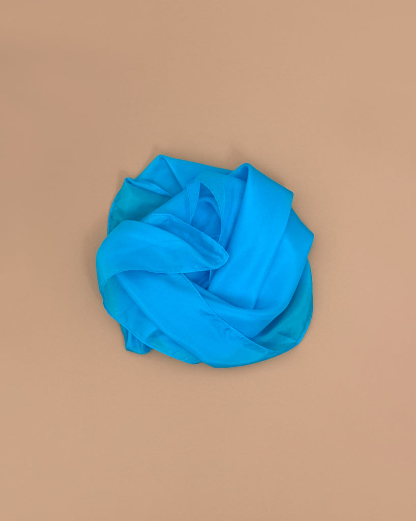 A vibrant turquoise Sarah's Silks Solid Color Playsilk is gently folded atop a neutral beige surface, forming soft curves for a simple, elegant look—ideal for open-ended play.