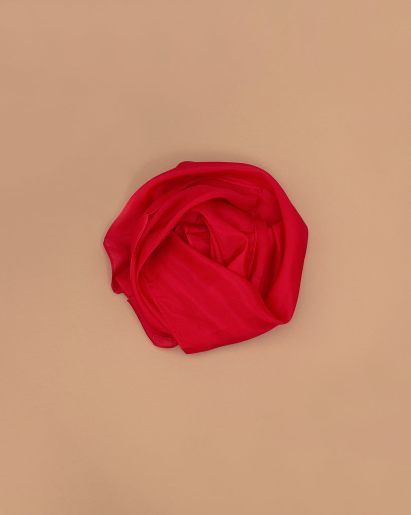 Sarah's Silks Solid Color Playsilk - Red, made of pure silk, is rolled into a circle and placed on a plain light brown background, inviting open-ended play.