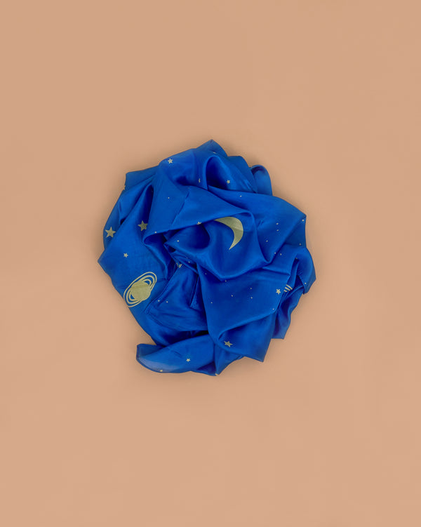 Sarah's Silks Enchanted Playsilk - Starry Night, a pure blue silk scarf with gold stars, moon, and planet designs, is loosely rolled on a beige background—ideal for open-ended play or as an enchanting playsilk accessory.