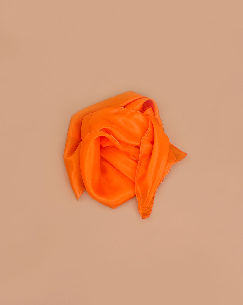 Sarah's Silks Solid Color Playsilk in Orange, loosely folded on a light brown background, inspires imaginative, open-ended play and brings elegance to sensory play activities.
