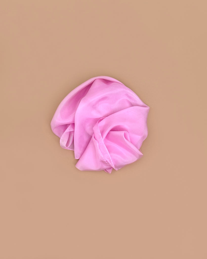 A Sarah's Silks Solid Color Playsilk in pink, made from pure silk and perfect for open-ended play, lies crumpled on a smooth, solid light brown background.