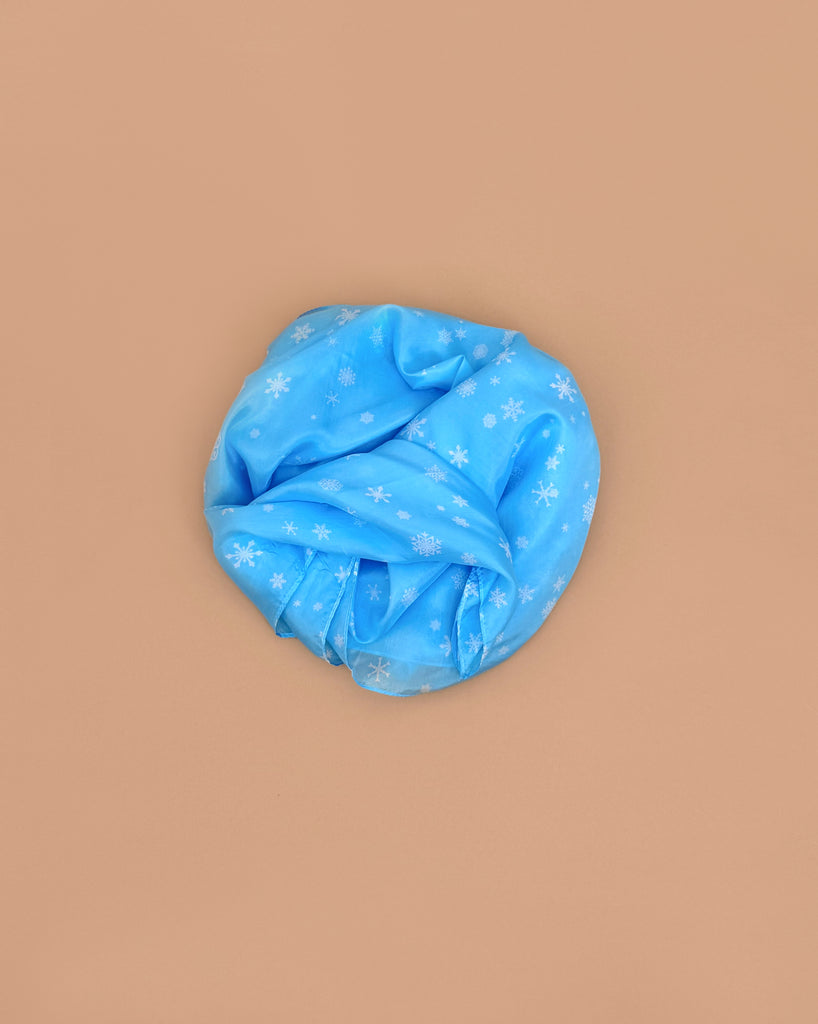A Sarah's Silks Giant Playsilk - Snow headwrap, light blue with white snowflakes, is displayed in a circle on a beige background, encouraging imaginative play inspired by Waldorf toys.