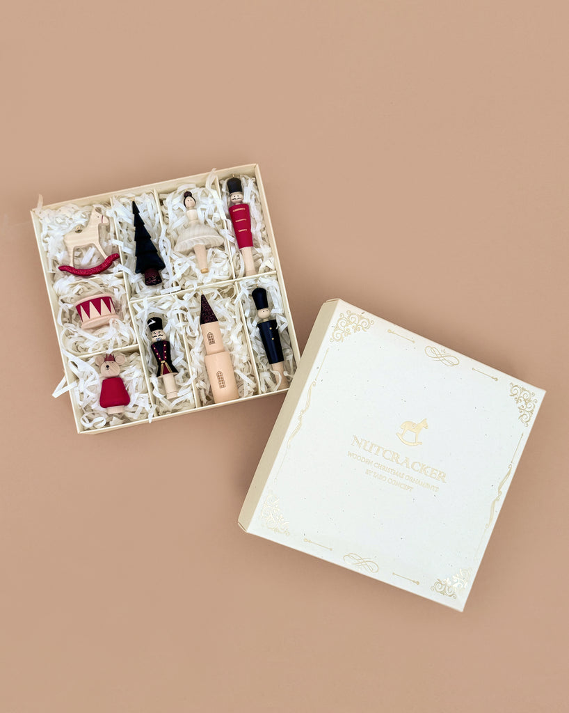 A cream Sabo Concept Christmas Ornaments box sits open on a tan surface, displaying nine hand-painted wooden Nutcracker-themed decorations—ballet dancers, a tree, and festive figures—nestled in shredded paper.