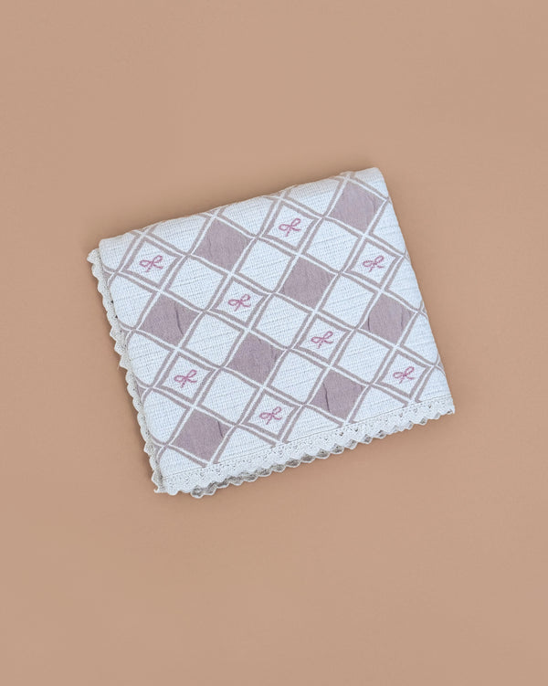The Organic Cotton Baby Patchwork Blanket - Pink Bows features a white base with pink and gray diamond patterns and small pink bows, displayed on a light brown background, evoking the charm of an embroidered keepsake.
