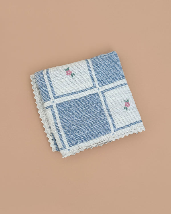 The Organic Cotton Patchwork Baby Blanket - Spring Bloom features blue and white checks with pink floral details, shown folded on a plain beige background.