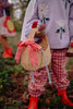 A child wearing pink checkered pants, a lavender sweater with flower designs, and red boots holds the Sticky Lemon Wicker Basket | Lemon Tree—a charming wicker accessory adorned with a red gingham bow. Another child stands nearby, partially visible.
