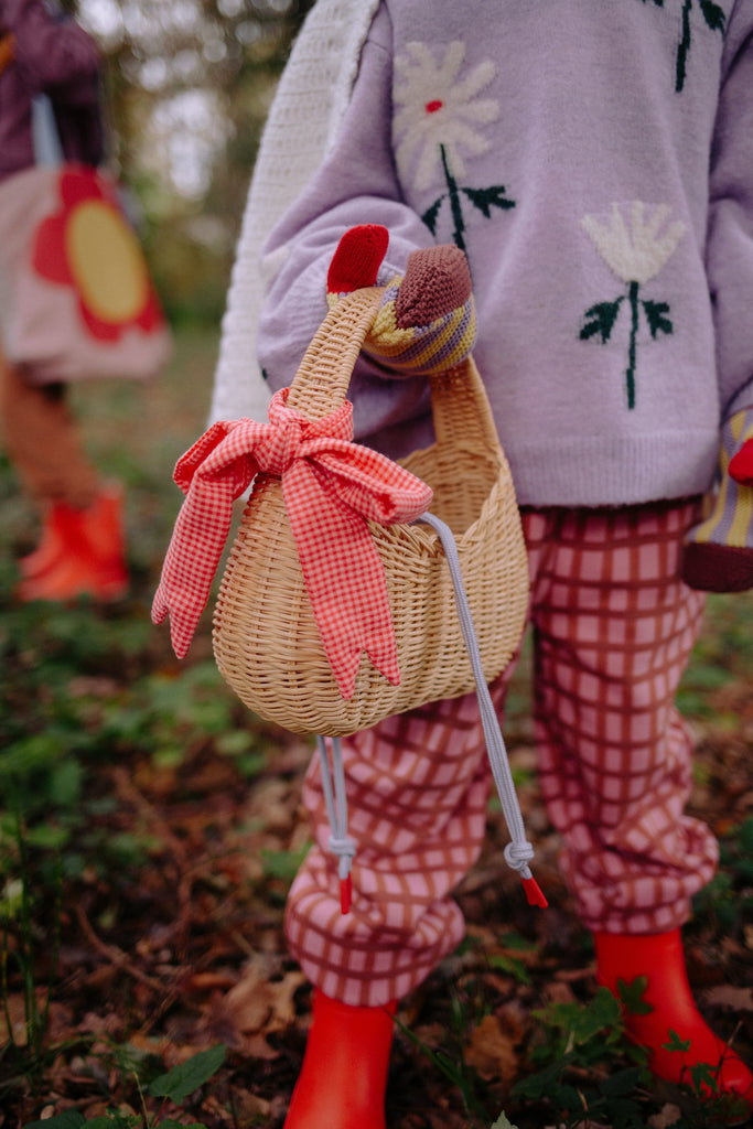 A child wearing pink checkered pants, a lavender sweater with flower designs, and red boots holds the Sticky Lemon Wicker Basket | Lemon Tree—a charming wicker accessory adorned with a red gingham bow. Another child stands nearby, partially visible.
