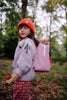 A young child wearing a red knit hat, patterned sweater, and the Sticky Lemon Backpack Small - Envelope - Rosebay Pink (ships in a week), featuring adjustable shoulder straps, stands outdoors in a forest, looking over their shoulder with a slight smile.
