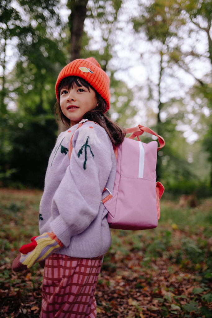 A young child wearing a red knit hat, patterned sweater, and the Sticky Lemon Backpack Small - Envelope - Rosebay Pink (ships in a week), featuring adjustable shoulder straps, stands outdoors in a forest, looking over their shoulder with a slight smile.