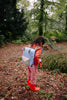 A child wearing a red raincoat and red boots, carrying the Sticky Lemon Backpack Medium | Envelope | Cloud Blue (ships in a week), stands on a leafy forest path, looking down at the ground, surrounded by green trees and foliage.
