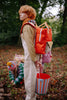 A person with reddish hair stands in a forest, wearing overalls and a scarf, and carrying the Sticky Lemon Backpack Medium | Envelope | Campfire Orange, which features waterproof lining and is made from recycled PET. A striped bucket hangs by their side. In the background, two children crouch on the ground gathering leaves.