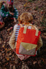 A child with a Sticky Lemon Backpack Medium | Envelope | Campfire Orange made from recycled PET, featuring bold red and yellow colors, crouches on leafy ground outdoors, facing away. In the background, another child in colorful clothing and a hat sits and reaches toward the ground.