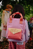A child with long dark hair stands outdoors wearing the Sticky Lemon Backpack Small – Envelope – Rosebay Pink, which features adjustable shoulder straps and is made from recycled PET. Two other children are blurred in the background among trees and leaves on the ground.