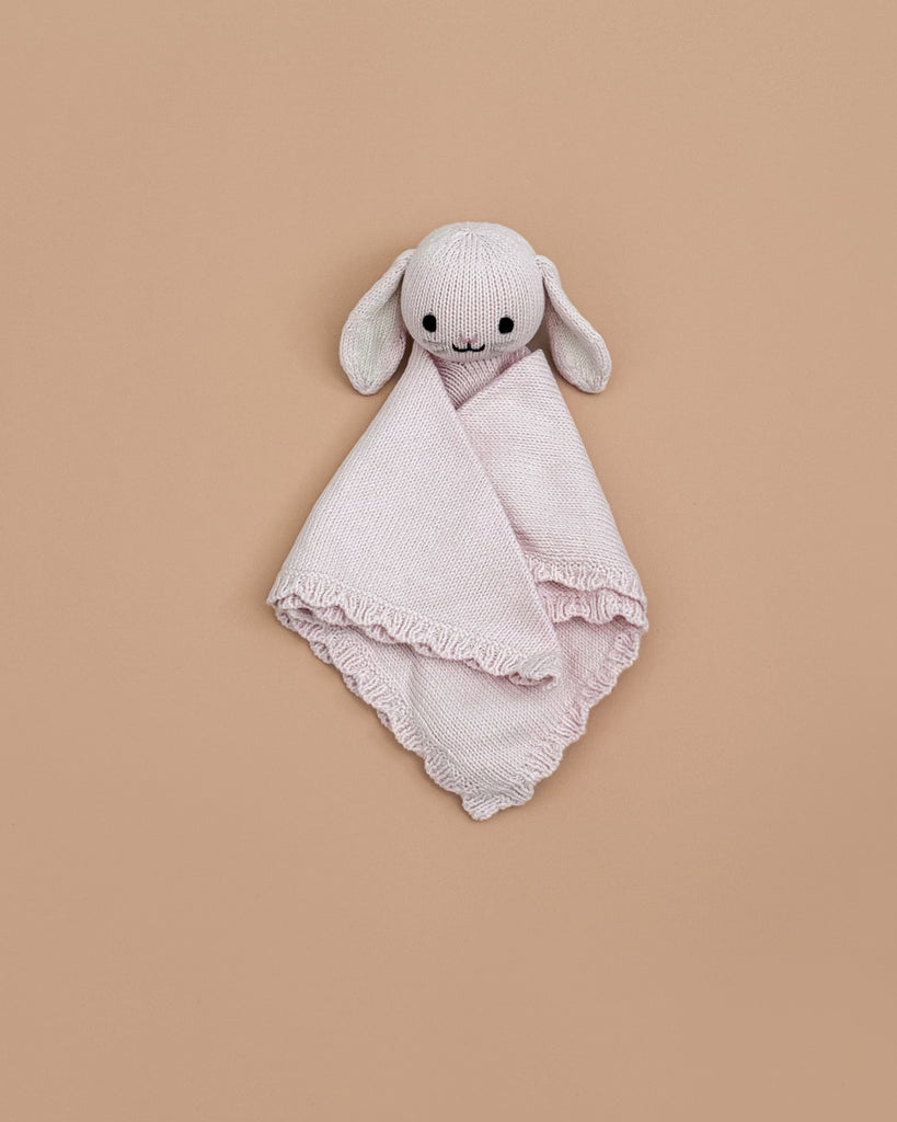 The Cuddle + Kind Organic Cotton Baby Bunny Lovey (Blush) is a soft, light pink heirloom-quality comforter with a cute bunny head and floppy ears, shown on a plain beige background.