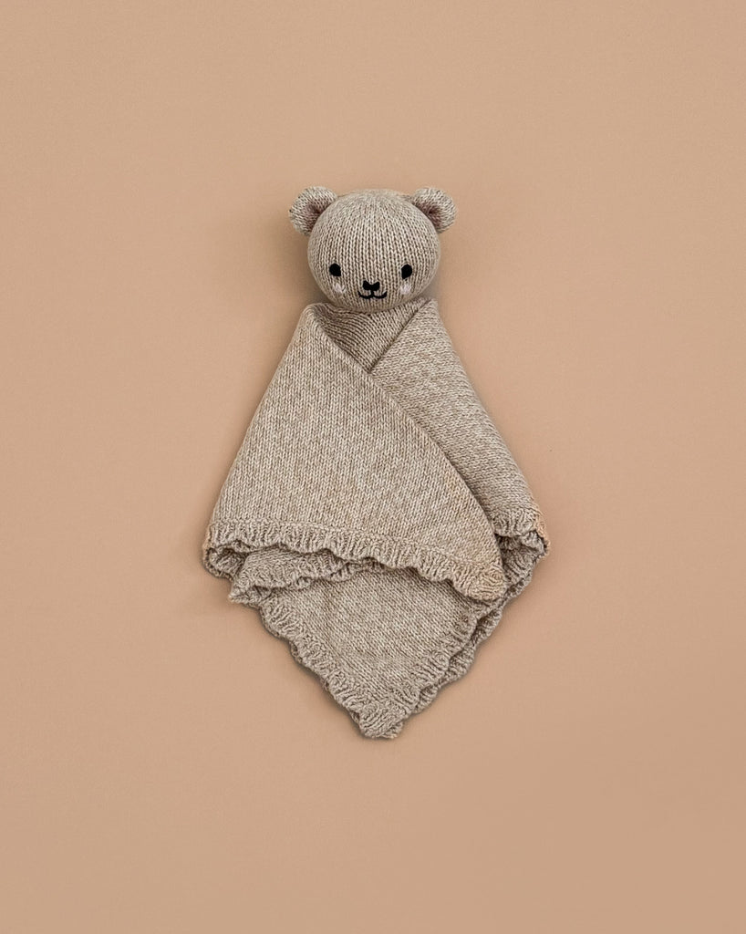 The Cuddle + Kind Organic Cotton Baby Bear Lovey (Honey) is a hand-knit beige teddy bear with round ears and a smiling face, wrapped in a soft, textured blanket against a light tan background.