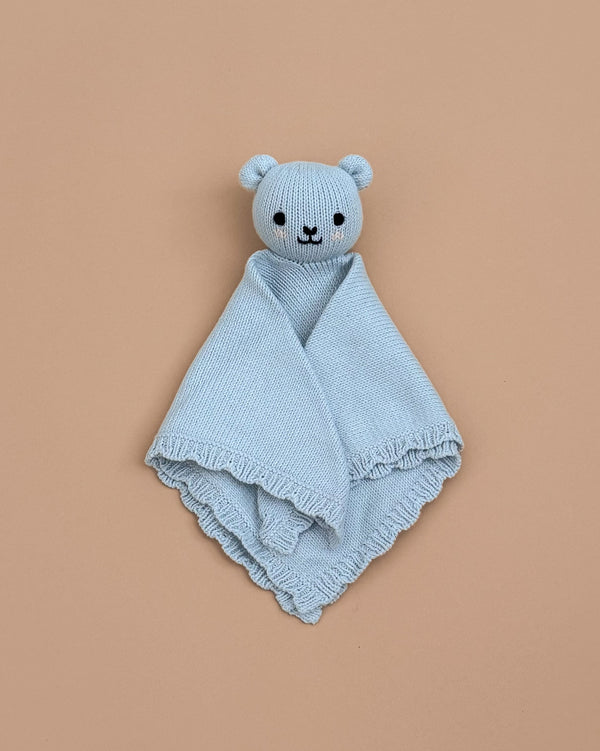The Cuddle + Kind Organic Cotton Baby Bear Lovey (Baby Blue) is hand-knit, featuring a bear’s head and arms with stitched eyes, nose, and smile, shown laid flat on a plain beige background.