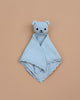 The Cuddle + Kind Organic Cotton Baby Bear Lovey (Baby Blue) is hand-knit, featuring a bear’s head and arms with stitched eyes, nose, and smile, shown laid flat on a plain beige background.
