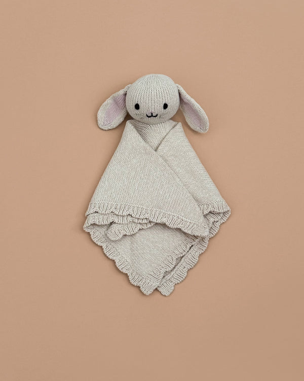 The Cuddle + Kind Organic Cotton Baby Bunny Lovey (Oatmeal), featuring floppy ears and embroidered facial details, is hand-knit and lies on a plain beige background.