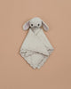 The Cuddle + Kind Organic Cotton Baby Bunny Lovey (Oatmeal), featuring floppy ears and embroidered facial details, is hand-knit and lies on a plain beige background.