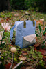 A Sticky Lemon Backpack Medium | Envelope | Cloud Blue (ships in a week), made from recycled PET bottles and featuring green straps, a beige front pocket, and a waterproof lining, sits on grass among artificial flowers and fallen leaves in an outdoor setting.