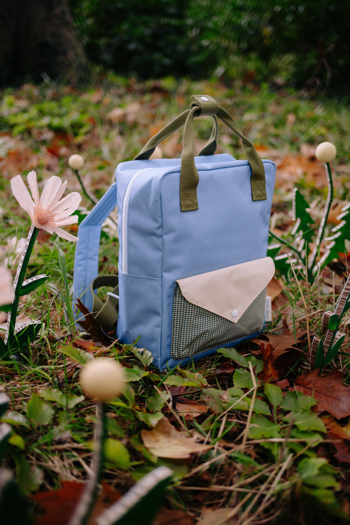 A Sticky Lemon Backpack Medium | Envelope | Cloud Blue (ships in a week), made from recycled PET bottles and featuring green straps, a beige front pocket, and a waterproof lining, sits on grass among artificial flowers and fallen leaves in an outdoor setting.