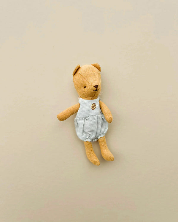 The Maileg Teddy Baby, dressed in a light blue and white gingham outfit, rests on a beige background. Its vintage charm and minimalistic design evoke nostalgia, reminiscent of days gone by.