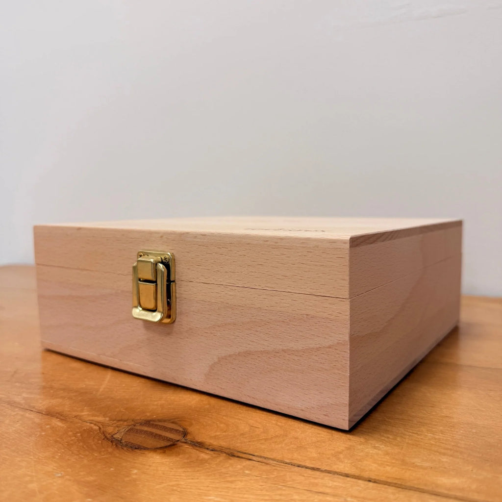 The Toverlux Silhouette Storage Box Deluxe, a light wooden box with a gold metal latch, sits on a wooden surface against a plain wall—perfect for organizing your cherished Toverlux Silhouettes in style.