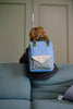 A child with light brown hair, wearing the Sticky Lemon Backpack Medium | Envelope | Cloud Blue—crafted from recycled PET bottles and featuring a blue design with a green handle—sits on a blue couch facing away from the camera against a light green wall.