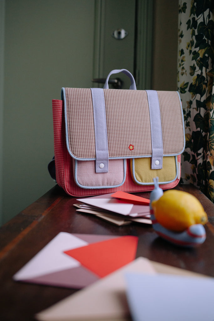 The Sticky Lemon School Bag | Lemon Tree | Gingham Flower + Rose, crafted from recycled PET bottles and accented with pastel pockets, rests on a wooden table surrounded by scattered envelopes and papers, with a lemon-shaped object close by. In the background, a green door and floral curtain add to the vibrant scene.