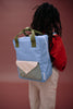 A person with long braided hair wears a red sweater and beige striped pants, carrying the Sticky Lemon Backpack Medium | Envelope in Cloud Blue—crafted from recycled PET bottles, featuring green handles and a front pocket—while standing against a maroon background.