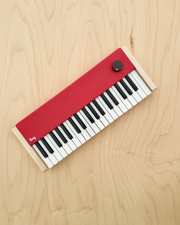 The Loog Piano is a compact, minimalist digital piano with velocity-sensitive black and white keys, a red panel, a single black knob, and light wooden sides, shown on a light wood surface.