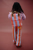 A person with long hair stands facing away, wearing the Sticky Lemon Backpack Medium | Stripes | Cloud Blue + Maple Whirl—featuring striped orange and blue design with waterproof lining made from recycled PET bottles—and matching pants, paired with a striped shirt, against a plain brown background.