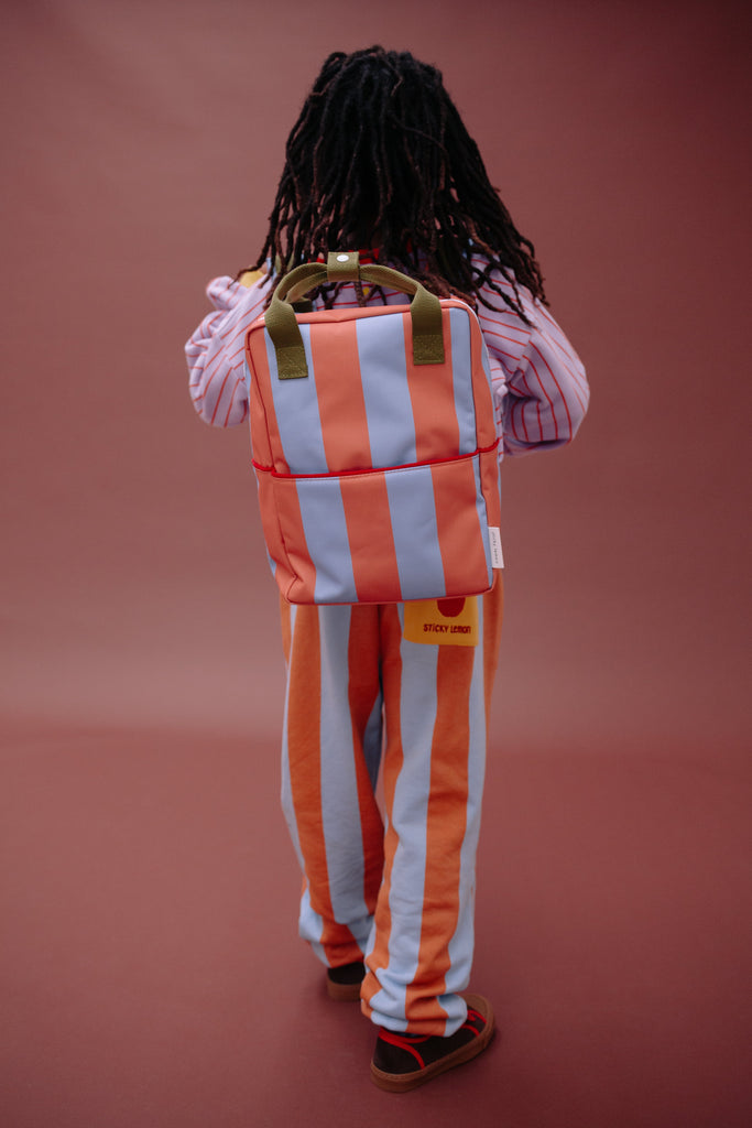 A person with long hair stands facing away, wearing the Sticky Lemon Backpack Medium | Stripes | Cloud Blue + Maple Whirl—featuring striped orange and blue design with waterproof lining made from recycled PET bottles—and matching pants, paired with a striped shirt, against a plain brown background.