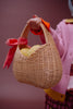 A person in a pink sweater holds the Sticky Lemon Wicker Basket | Lemon Tree, adorned with a red bow and filled with yellow fruit, against a plain background.