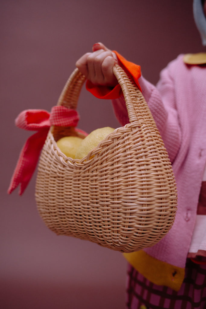 A person in a pink sweater holds the Sticky Lemon Wicker Basket | Lemon Tree, adorned with a red bow and filled with yellow fruit, against a plain background.